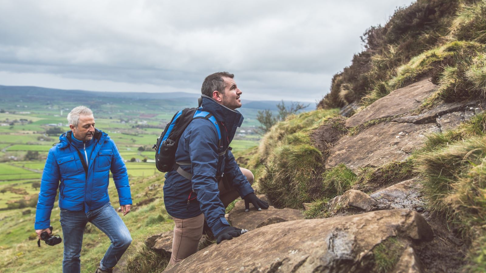 Two men climbing up Slemish Mountain with views of the countryside behind